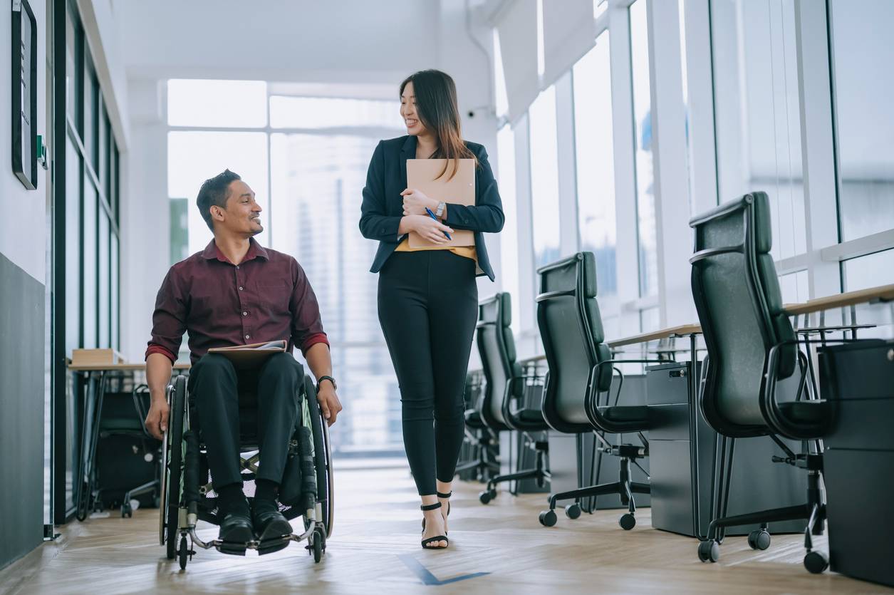 Indian white collar male worker in wheelchair having cheerful discussion conversation with his female asain chinese colleague coworking at walkway corridor stock photo